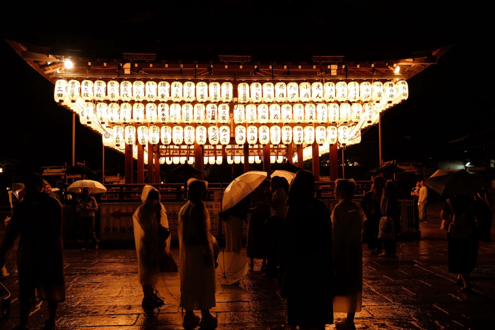 Yasaka Shrine's stage in the rain at night, rows of paper lanterns above people with clear umbrellas